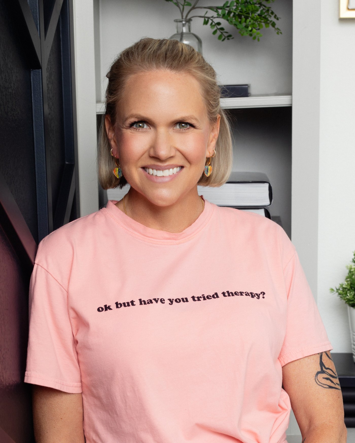 SarahBeth Bolin, LCSW, smiling at the camera in a t-shirt printed with the words “ok but have you tried therapy?”, wearing rainbow heart earrings.